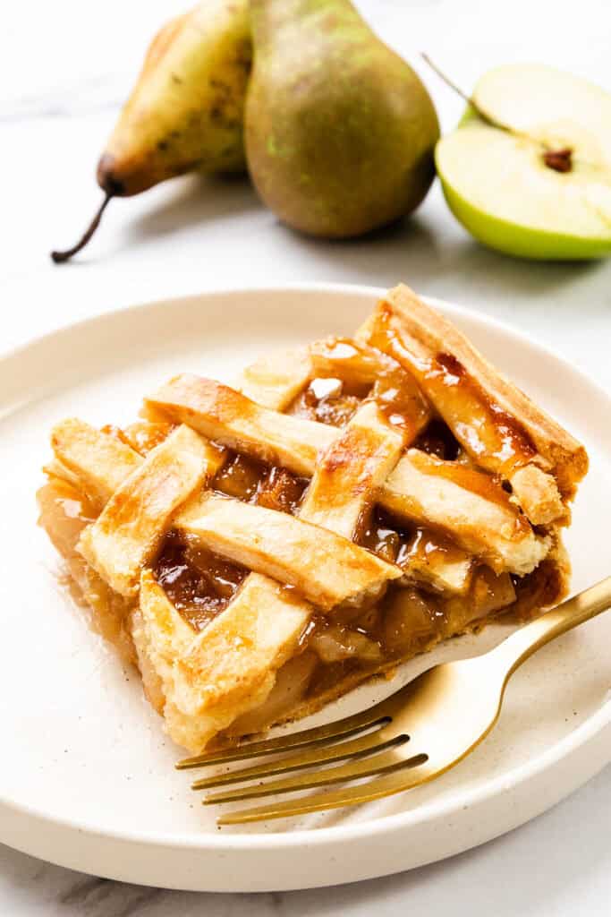 A slice of lattice-topped pear pie on a white plate, with a gold fork beside it. Whole and sliced pears enhance the scene on a white countertop, resembling a delectable apple pie presentation.