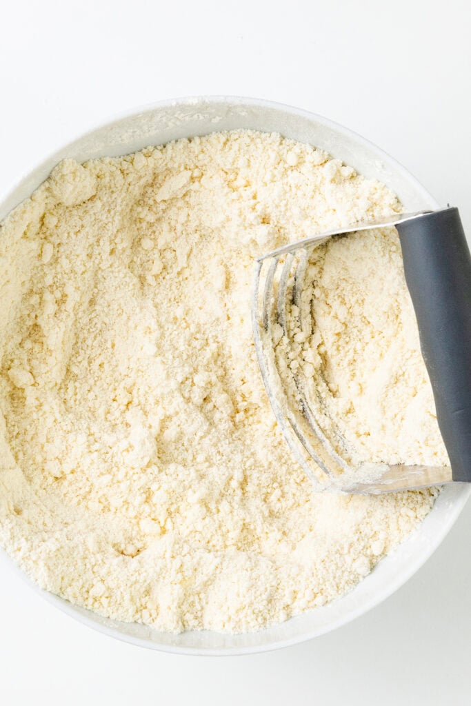 A bowl filled with flour and a pastry cutter resting on top, perfect for crafting a delicious pear pie. The flour appears finely sifted, and the pastry cutter has a black handle. The image is taken from above on a white background.