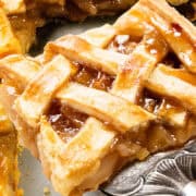 A close-up of a slice of apple pie on a serving spatula. The golden-brown, lattice-topped crust is flaky, with a shiny glaze and visible apple filling. The pie rests on a gray surface, alongside another partial slice tantalizingly peeking from behind.