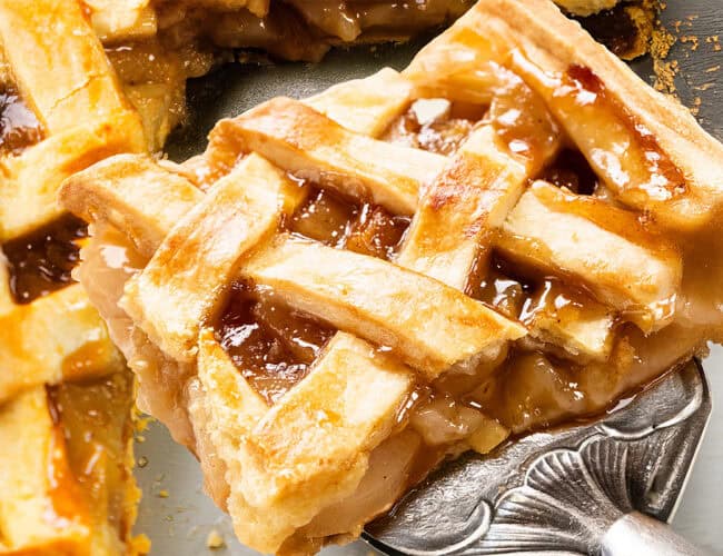 A close-up of a slice of apple pie on a serving spatula. The golden-brown, lattice-topped crust is flaky, with a shiny glaze and visible apple filling. The pie rests on a gray surface, alongside another partial slice tantalizingly peeking from behind.