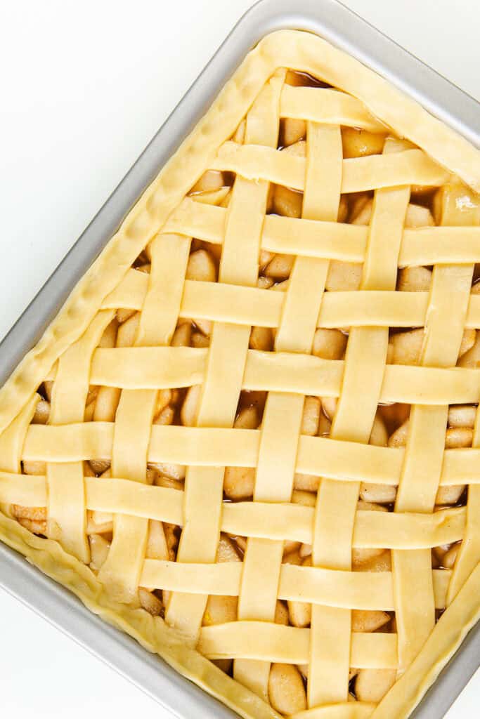 A rectangular pan holds an unbaked apple pie with a lattice crust. The dough is expertly woven in a crisscross pattern over the succulent apple filling. This delightful creation resembles an inviting pear pie, ready for the oven, and rests gracefully on a white surface.