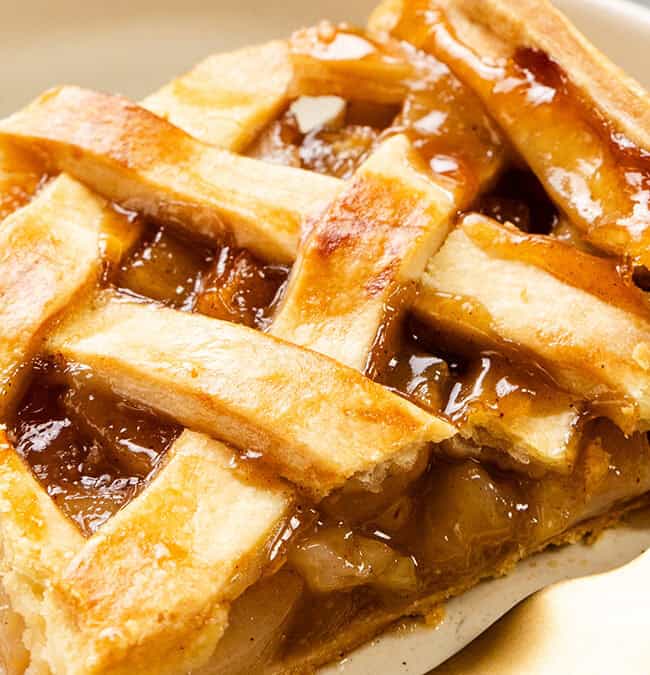 A close-up of a slice of apple pie with a golden-brown lattice crust on a white plate. A gold fork is placed beside the pie, and the filling appears glossy and caramelized.