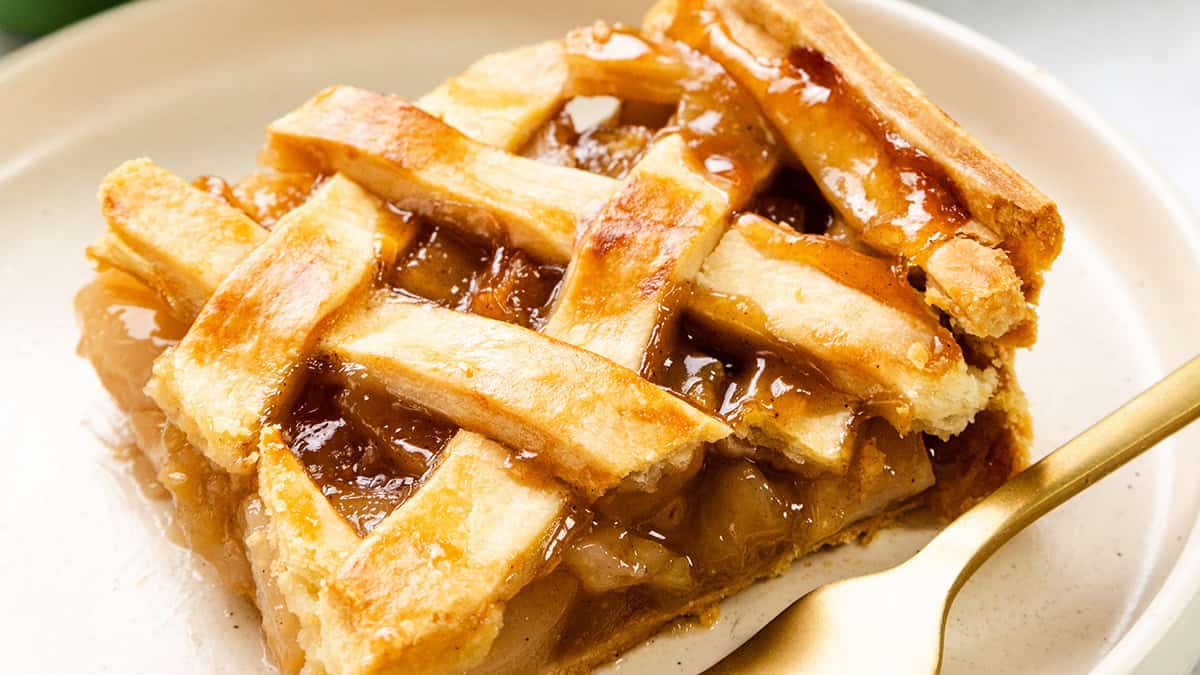 A close-up of a slice of apple pie with a golden-brown lattice crust on a white plate. A gold fork is placed beside the pie, and the filling appears glossy and caramelized.