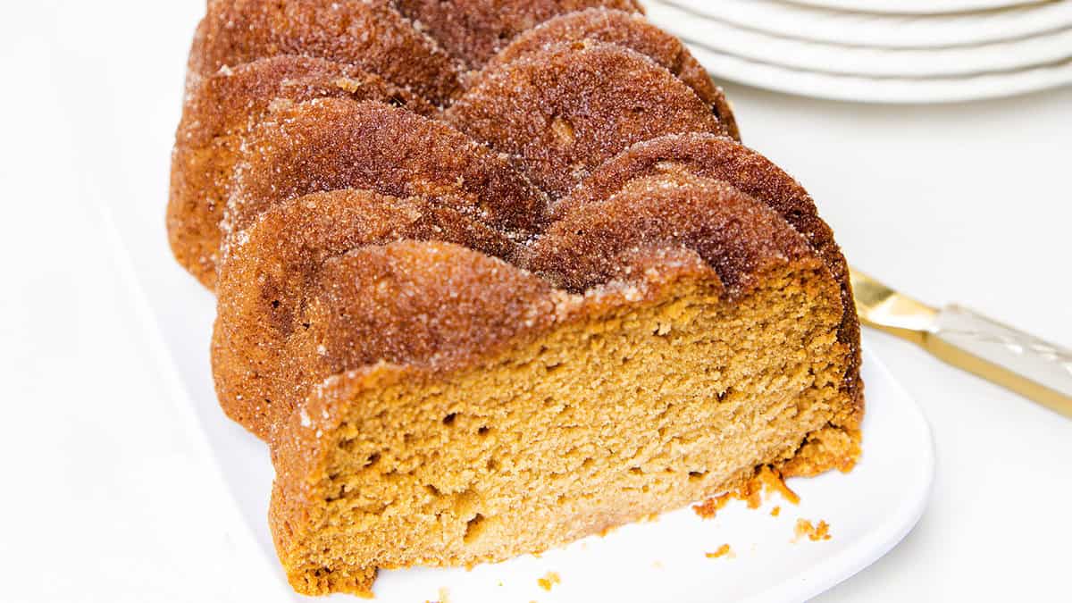 A close-up of a sliced brown sugar pound cake on a white plate. The cake has a textured, golden-brown crust and a moist interior. A gold-handled knife is beside the plate, and white dishes are stacked in the background.