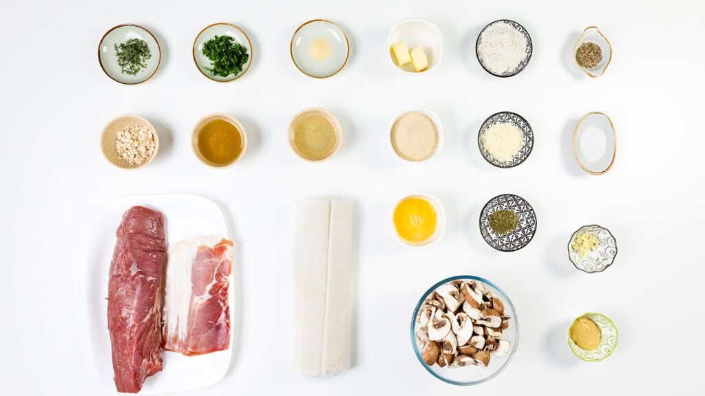 The ingredients for Beef Wellington are neatly arranged on a pristine white background, featuring raw beef, prosciutto, puff pastry, chopped herbs, mushrooms, eggs, butter, cream, and small bowls of spices and seasonings.