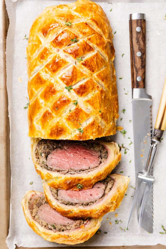 A beautifully sliced Beef Wellington rests on a parchment-lined tray, showcasing tender beef wrapped in golden puff pastry and flavorful mushroom duxelles. A knife and fork lie beside it, perfectly garnished with fresh thyme.