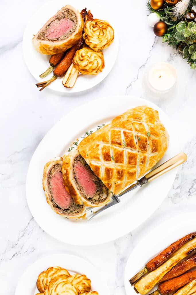 A beautifully sliced Beef Wellington rests on a white platter, encircled by roasted carrots and duchess potatoes. In the background, soft candlelight and a decorative wreath add a festive touch to the scene.