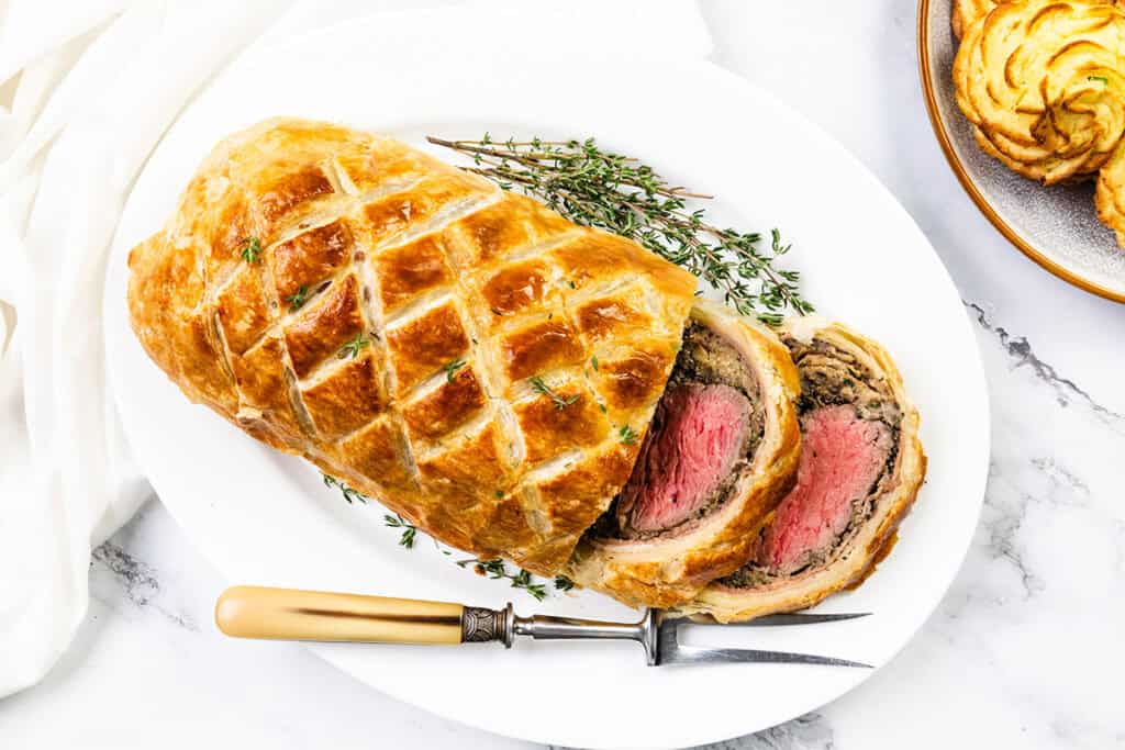 The succulent Beef Wellington, enveloped in a golden, flaky pastry crust, is sliced to reveal its tender, pink center. Garnished with fresh thyme, it sits elegantly on a white plate beside a vintage knife with a light handle.