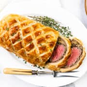 A succulent beef Wellington graces a white plate, garnished with fresh thyme. The golden brown pastry is crosshatched beautifully, revealing perfectly pink beef inside. A carving knife with a wooden handle rests elegantly beside it on the marble surface.