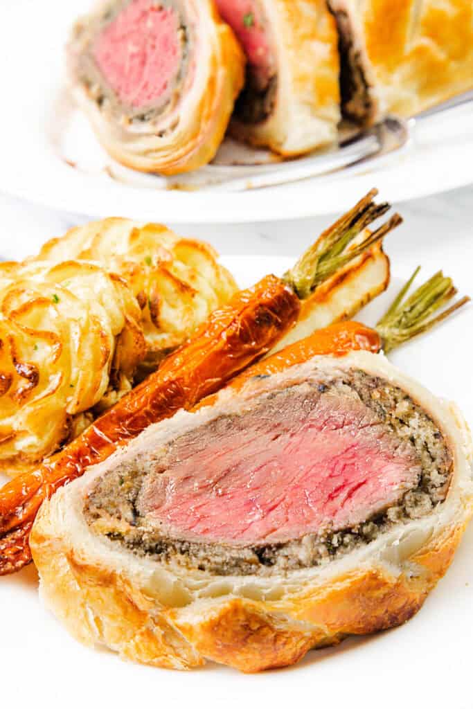 A close-up of Beef Wellington showcases its golden puff pastry crust, sliced to reveal pink, tender meat inside. This culinary masterpiece is perfectly paired with roasted carrots and crispy potato rosettes on a pristine white plate.