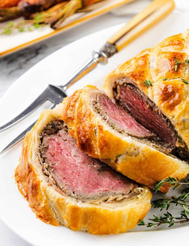 A close-up of Beef Wellington reveals medium-rare beef encased in mushroom duxelles and golden, flaky pastry. Garnished with fresh thyme, roasted carrots sit in the background on a white plate.