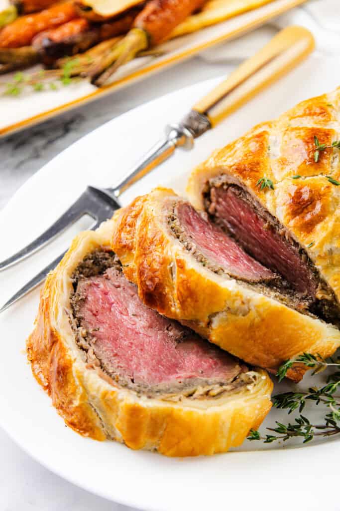 A close-up of Beef Wellington reveals medium-rare beef encased in mushroom duxelles and golden, flaky pastry. Garnished with fresh thyme, roasted carrots sit in the background on a white plate.