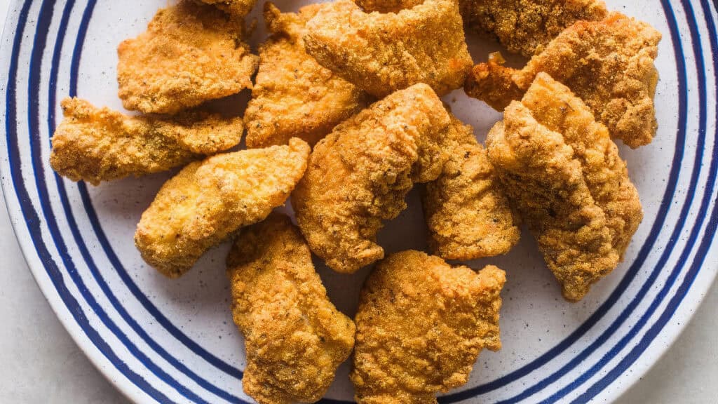 A plate of crispy, golden fried catfish pieces, inspired by Southern recipes, is arranged on a white dish with blue stripes.