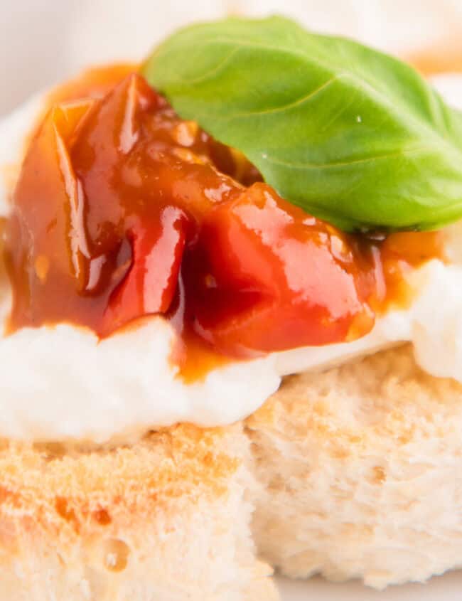 A slice of bread topped with a dollop of creamy cheese, a spoonful of tomato jam, and a fresh basil leaf makes for perfect Christmas party food. The dish sits on a white plate, its festive appeal enhanced by another similar piece blurred in the background.