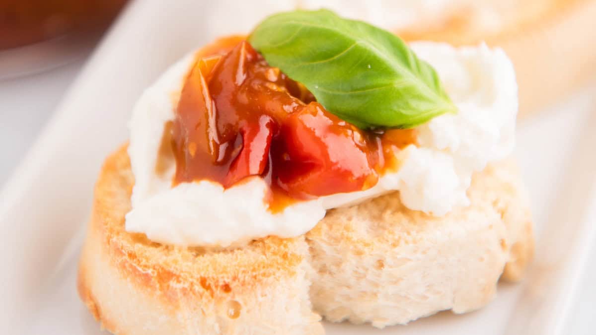 A slice of bread topped with a dollop of creamy cheese, a spoonful of tomato jam, and a fresh basil leaf makes for perfect Christmas party food. The dish sits on a white plate, its festive appeal enhanced by another similar piece blurred in the background.