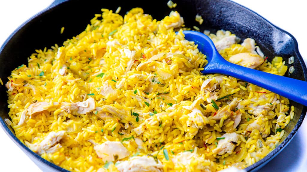 A skillet filled with yellow rice mixed with shredded chicken and garnished with herbs—a tried-and-true recipe that's sure to be a hit. A blue spoon rests on the side of the pan.