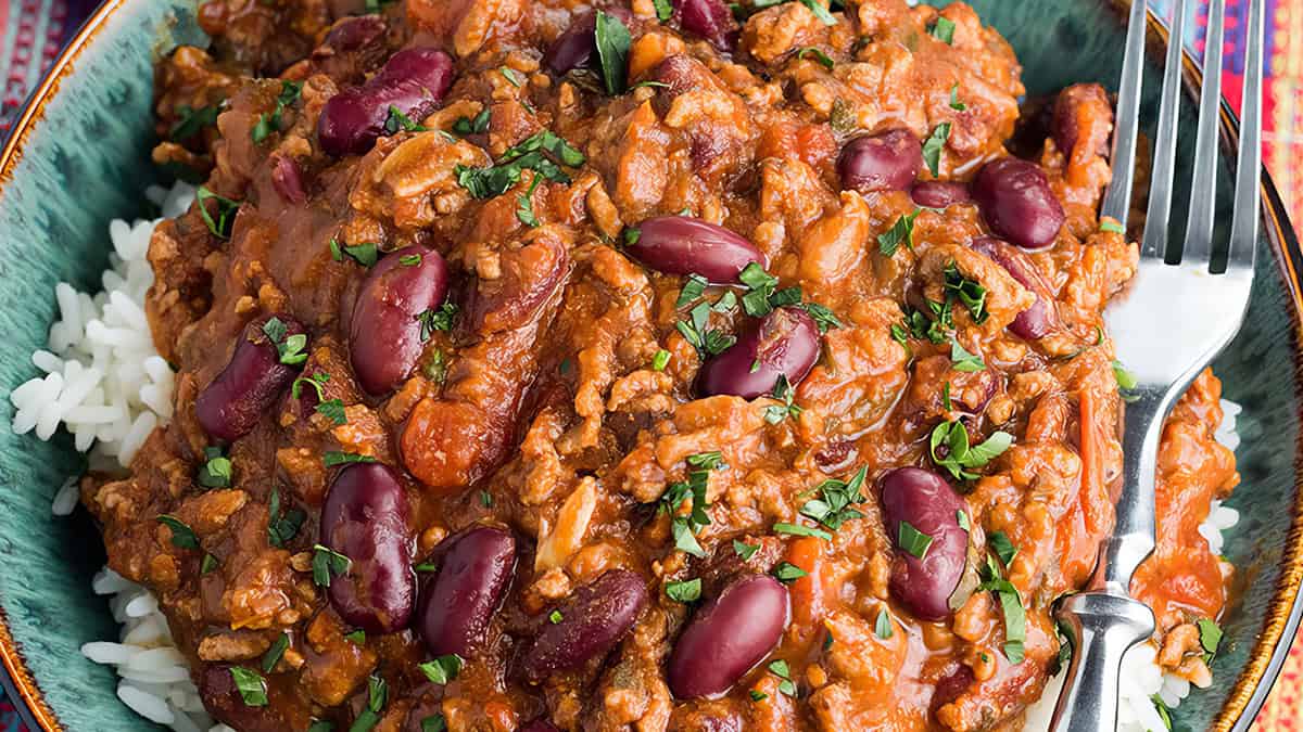 A bowl of chili con carne served over white rice, garnished with chopped herbs. The dish includes kidney beans and ground meat in a rich, red sauce. A fork is placed beside the chili in the bowl.