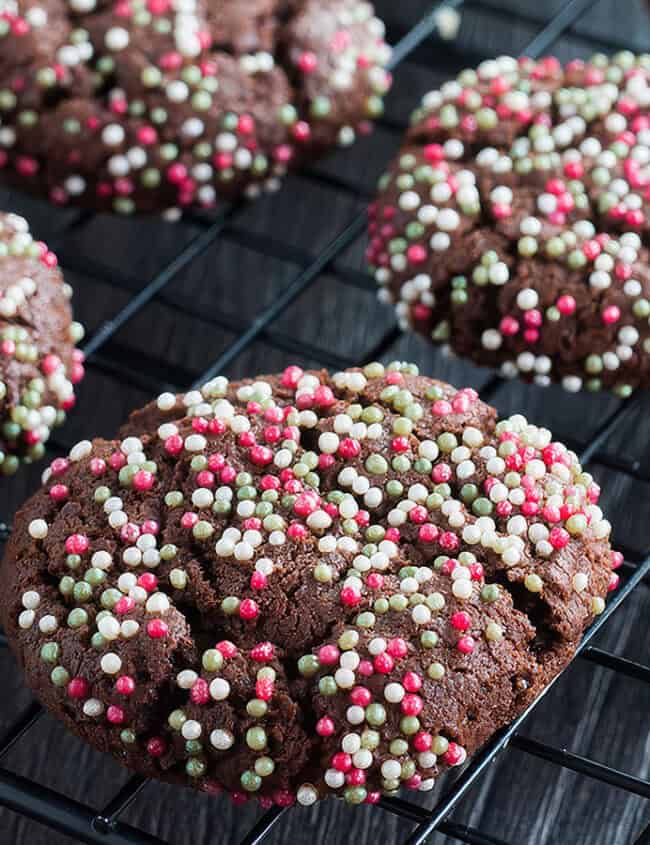 Chocolate cookies covered with red, white, and green sprinkles are cooling on a black wire rack. The background is a dark surface.