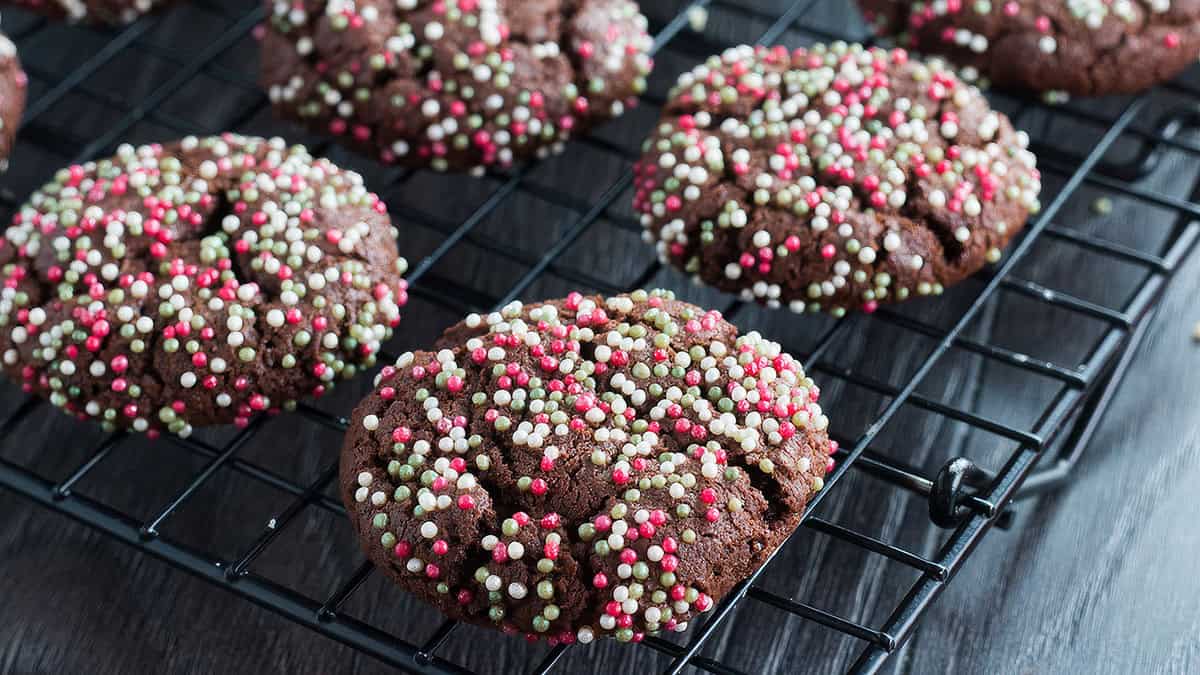 Chocolate cookies covered with red, white, and green sprinkles are cooling on a black wire rack. The background is a dark surface.