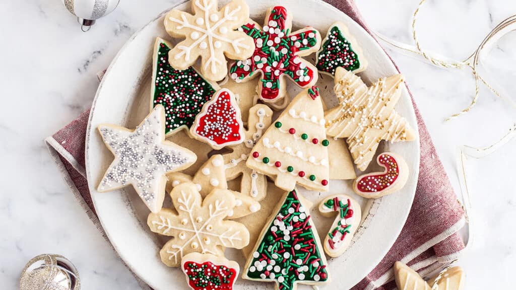 A plate of festive Christmas cookies shaped like trees, stars, snowflakes, and candy canes graces the table. These delightful Christmas cookies are decorated with colorful icing and sprinkles, set beautifully on a red and white napkin.