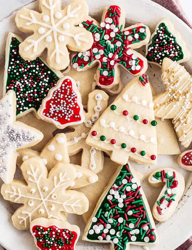 A plate of festive Christmas cookies shaped like trees, stars, snowflakes, and candy canes graces the table. These delightful Christmas cookies are decorated with colorful icing and sprinkles, set beautifully on a red and white napkin.