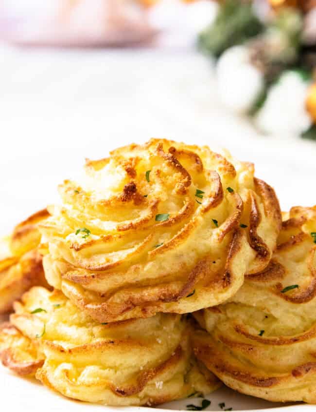 A close-up of golden brown duchess potatoes, intricately piped like a gourmet mashed potatoes recipe, and garnished with chopped herbs on a white plate. The background features a blurred festive setting.