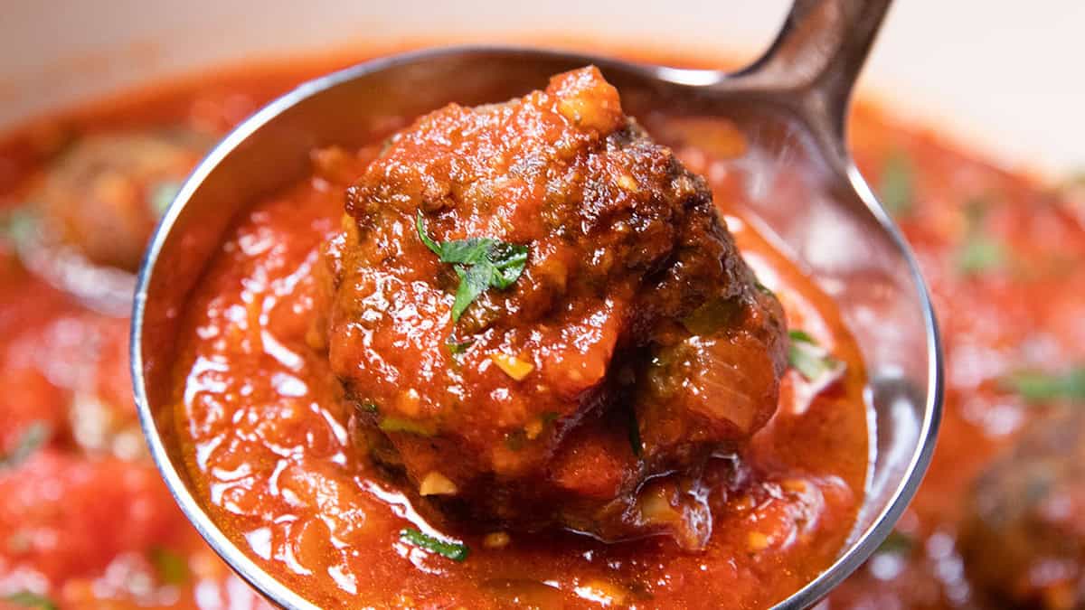 A close-up of a ladle holding a meatball covered in rich tomato sauce with a sprinkle of herbs. The background shows more meatballs in the sauce, suggesting a warm, hearty dish.