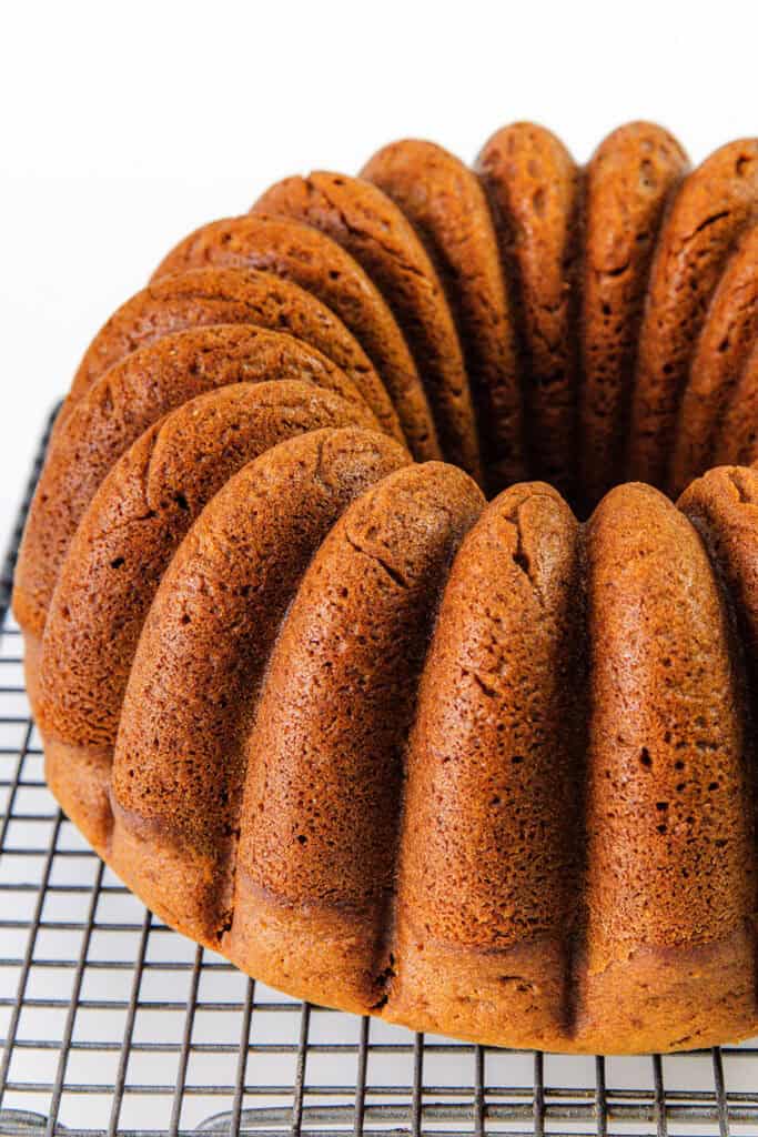 A golden-brown gingerbread bundt cake with a scalloped design sits on a metal cooling rack. The cake's smooth and even texture is visually appealing against the white background.