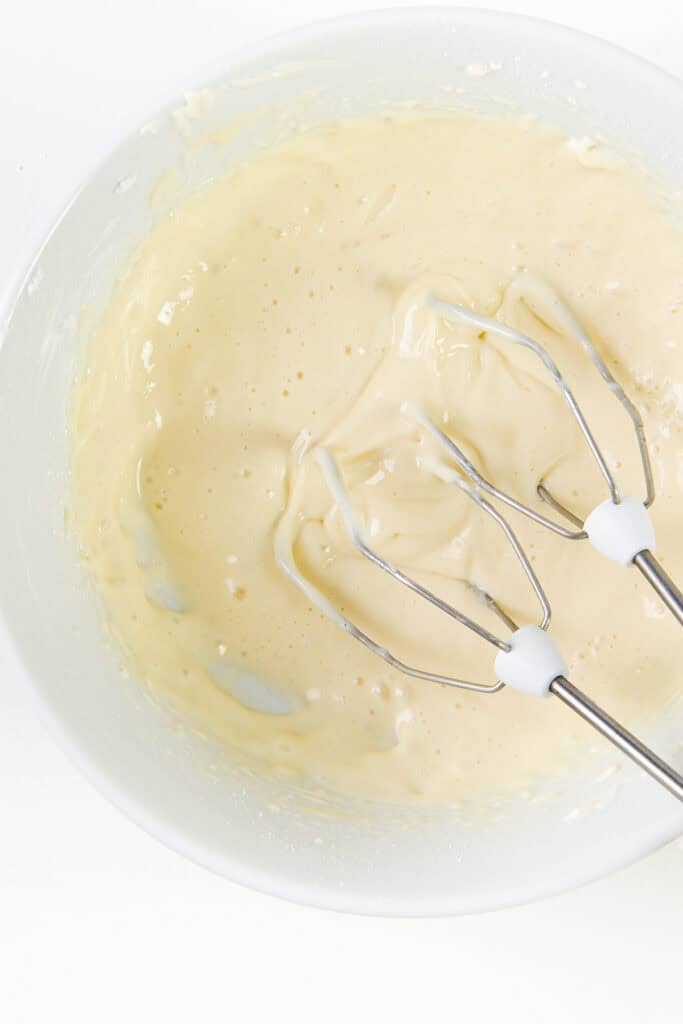 A close-up of a mixing bowl with creamy gingerbread batter. Two beaters from an electric mixer are resting in the mixture, which is pale yellow with a smooth and slightly fluffy texture, hinting at the perfect cake consistency.