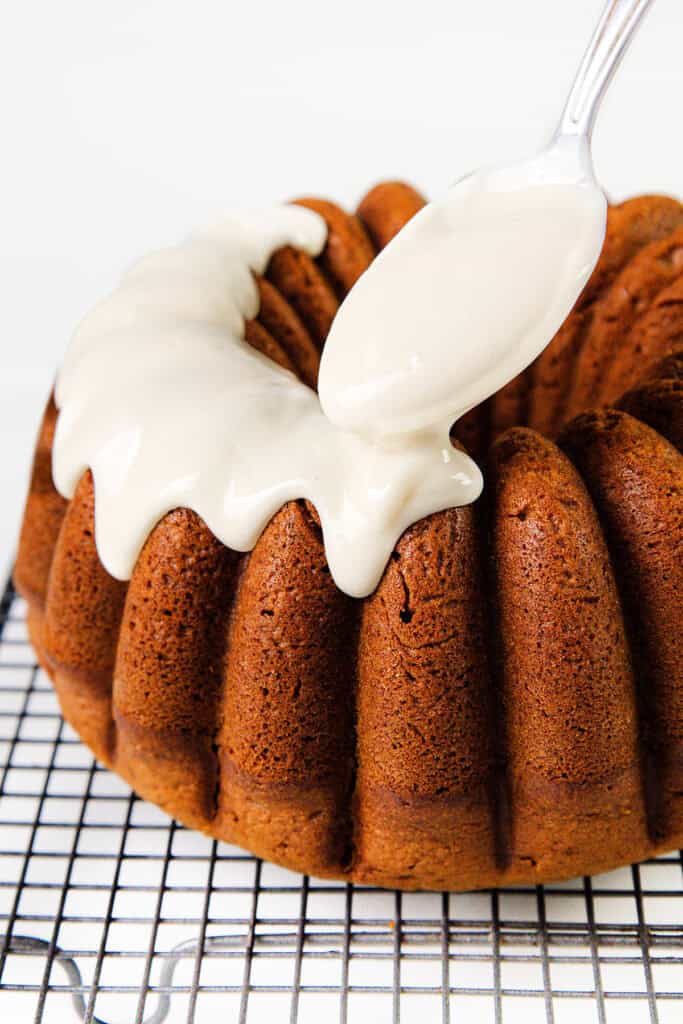 A gingerbread Bundt cake rests on a cooling rack as a spoonful of creamy, white icing is artfully draped over its ridged edges, providing a smooth contrast to the rich brown beneath.