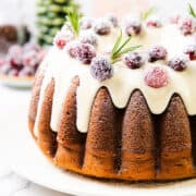 A festive gingerbread bundt cake topped with white icing, garnished with sugared cranberries and rosemary sprigs. The background features a blurred Christmas tree and decorative items, adding to the holiday charm.