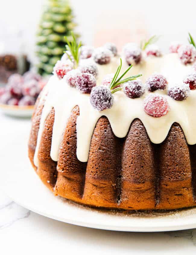 A festive gingerbread bundt cake topped with white icing, garnished with sugared cranberries and rosemary sprigs. The background features a blurred Christmas tree and decorative items, adding to the holiday charm.