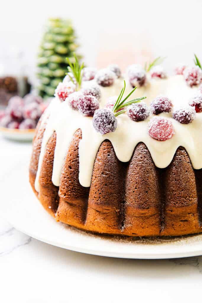 A festive gingerbread bundt cake topped with white icing, garnished with sugared cranberries and rosemary sprigs. The background features a blurred Christmas tree and decorative items, adding to the holiday charm.