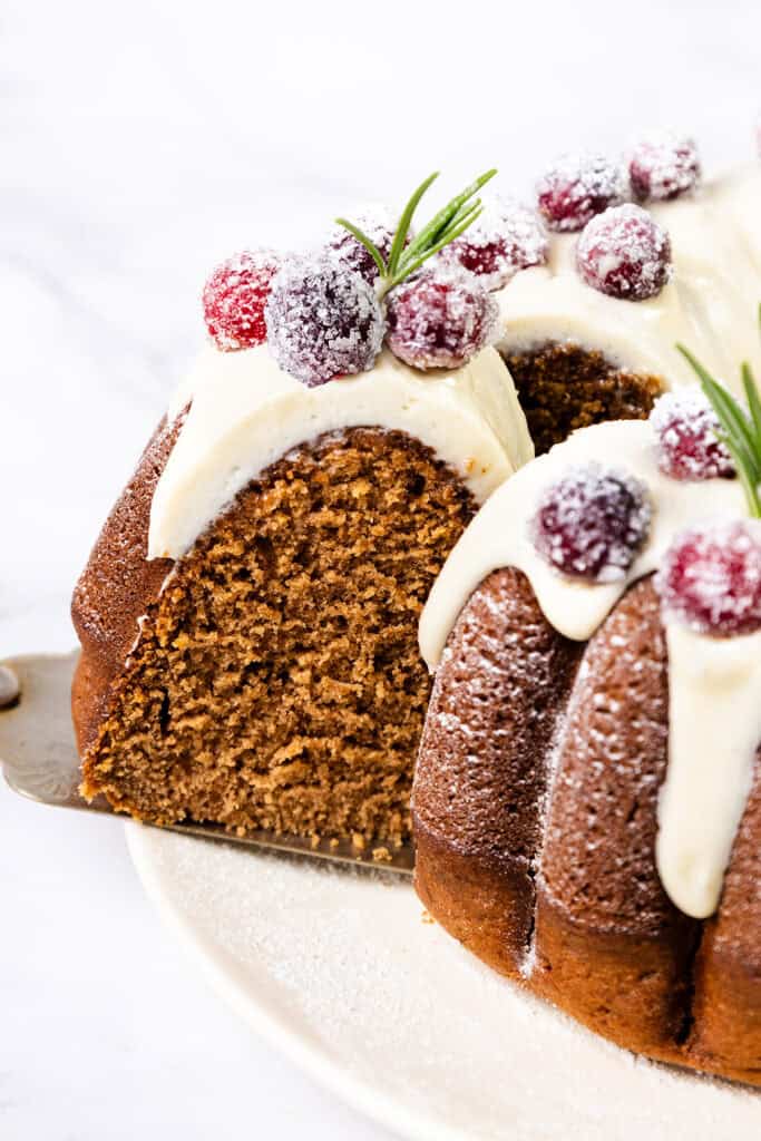 A close-up of a Bundt cake topped with white icing and sugared cranberries. A slice is being taken out, revealing the moist gingerbread interior. The cake is garnished with rosemary sprigs on a white plate.