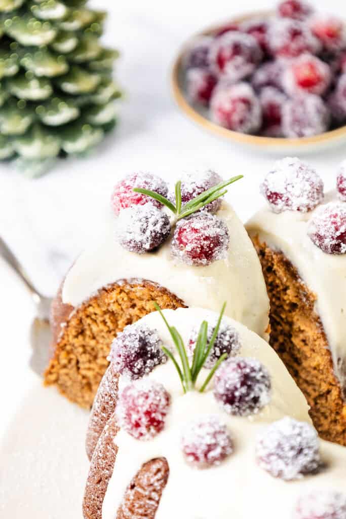 A gingerbread bundt cake topped with creamy icing and sugared cranberries, accented by a sprig of rosemary for a festive touch. In the background, a small dish holds more sugared cranberries, while a mini evergreen tree peeks into the scene.