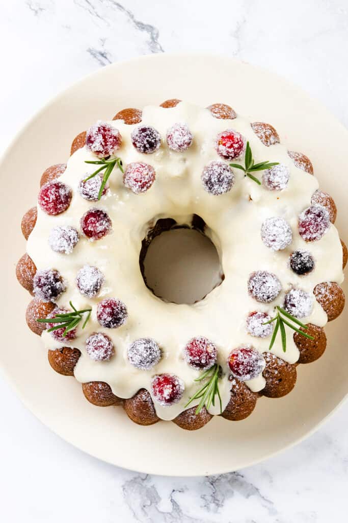 A festive gingerbread Bundt cake topped with white icing, sugared cranberries, and sprigs of rosemary graces a white plate. The cake is set against a light marble background, exuding seasonal charm.