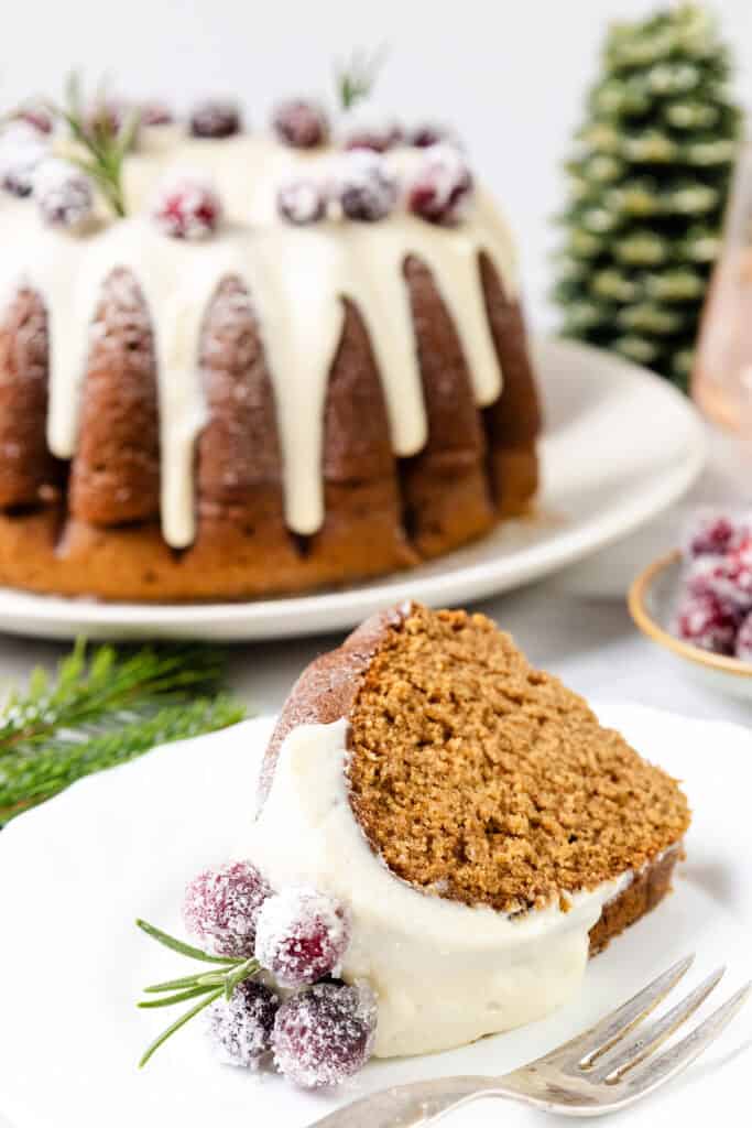 A slice of gingerbread bundt cake with white icing is served on a plate, capturing the warmth of holiday flavors. In the background, the gingerbread bundt cake is elegantly adorned with sugared cranberries and rosemary sprigs, while a small evergreen tree stands nearby.