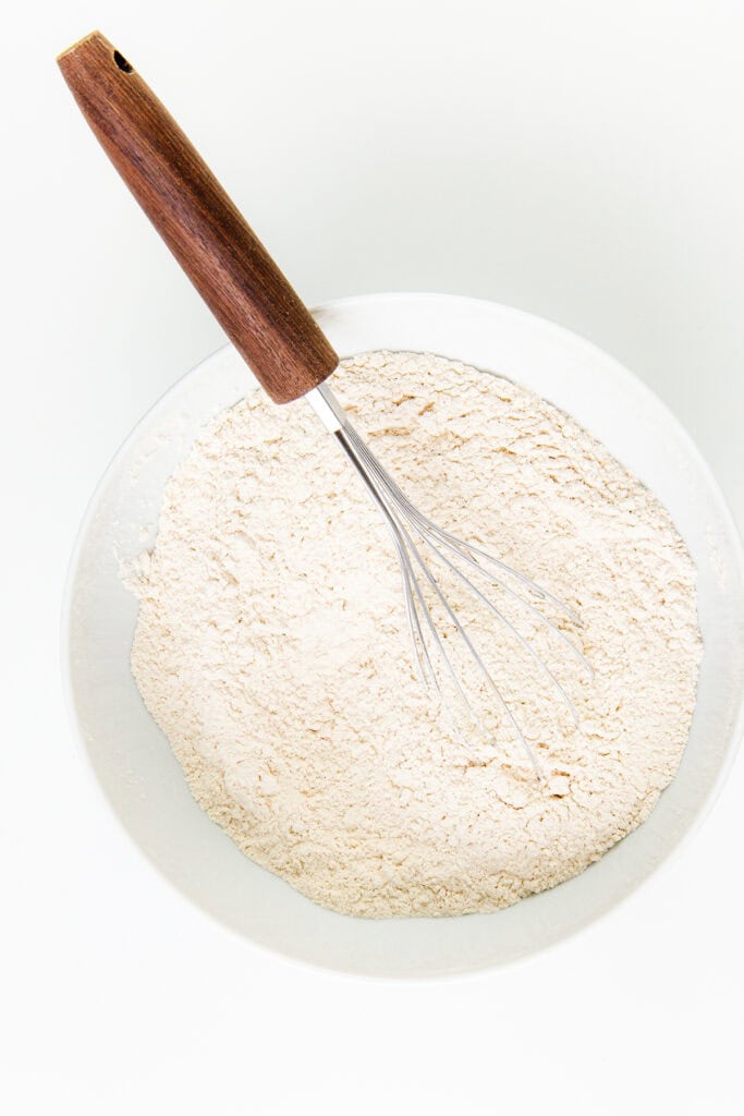 A mixing bowl filled with a mound of flour and a whisk resting inside, ready to craft a perfect cake. The bowl sits on a white surface, and the whisk, with its wooden handle, awaits your gingerbread masterpiece.