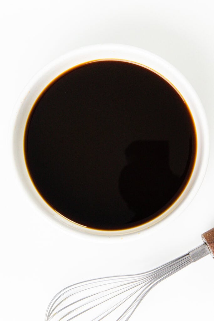 A white bowl filled with dark soy sauce sits on a white surface, reminiscent of the rich glaze on a gingerbread cake. A silver whisk with a wooden handle lies beside the bowl, ready to blend flavors seamlessly.