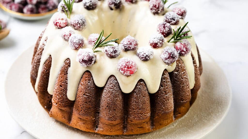 A Gingerbread Bundt cake topped with white icing, sugared cranberries, and sprigs of rosemary rests on a white plate. Set on a marble surface, it presents an elegant and festive appearance.