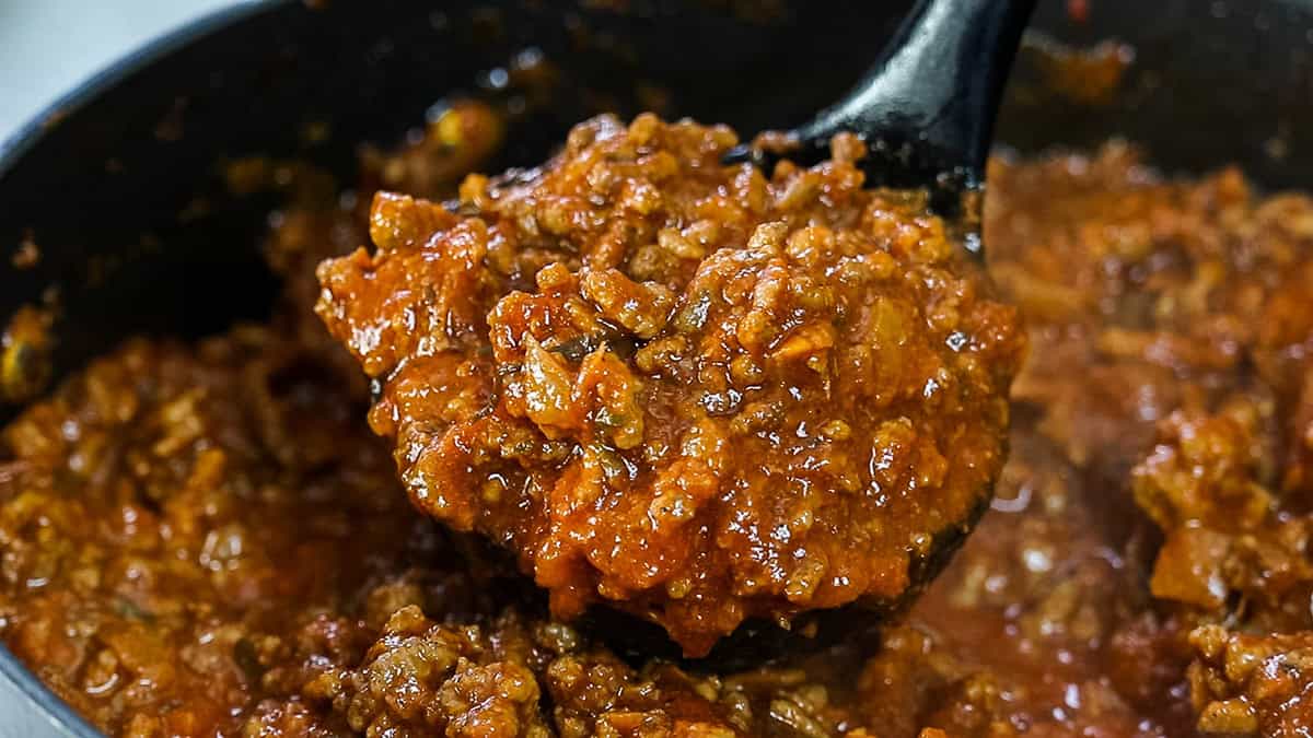 A close-up view of a ladle scooping rich, reddish-brown meat sauce from a pot. The sauce is chunky and well-cooked, with visible pieces of ground meat and tomato, suggesting a hearty texture and flavorful dish.