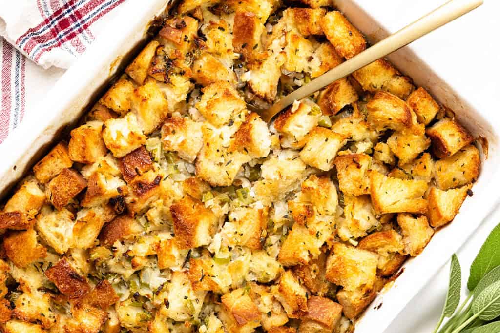 A close-up of make-ahead herb stuffing in a rectangular dish. The stuffing is golden brown with visible chunks of bread, herbs, and chopped celery. A spoon rests in the dish, ready for serving. A red and white striped cloth is nearby.