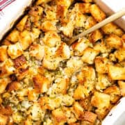 A close-up of make-ahead herb stuffing in a rectangular dish. The stuffing is golden brown with visible chunks of bread, herbs, and chopped celery. A spoon rests in the dish, ready for serving. A red and white striped cloth is nearby.