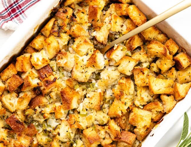 A close-up of make-ahead herb stuffing in a rectangular dish. The stuffing is golden brown with visible chunks of bread, herbs, and chopped celery. A spoon rests in the dish, ready for serving. A red and white striped cloth is nearby.