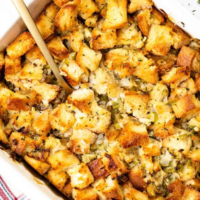 A casserole dish brimming with golden-brown herb stuffing, featuring chopped celery and onions. A wooden spoon rests invitingly in the dish, ready to serve. A red-striped towel peeks into the frame, adding a touch of charm to this make-ahead stuffing delight.