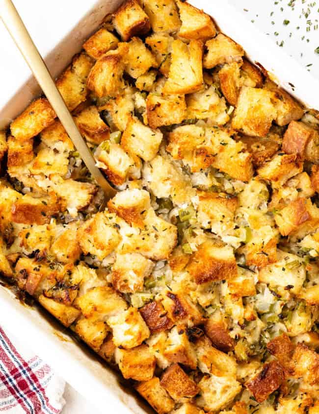 A casserole dish brimming with golden-brown herb stuffing, featuring chopped celery and onions. A wooden spoon rests invitingly in the dish, ready to serve. A red-striped towel peeks into the frame, adding a touch of charm to this make-ahead stuffing delight.