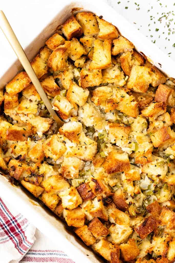 A casserole dish brimming with golden-brown herb stuffing, featuring chopped celery and onions. A wooden spoon rests invitingly in the dish, ready to serve. A red-striped towel peeks into the frame, adding a touch of charm to this make-ahead stuffing delight.