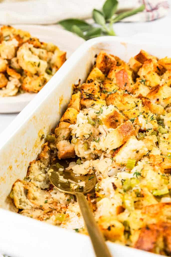 A casserole dish filled with Thanksgiving Stuffing, showcasing chunks of bread, celery, and herbs. A gold spoon rests inside the dish, while a separate plate with a serving of the herb stuffing is visible in the background.