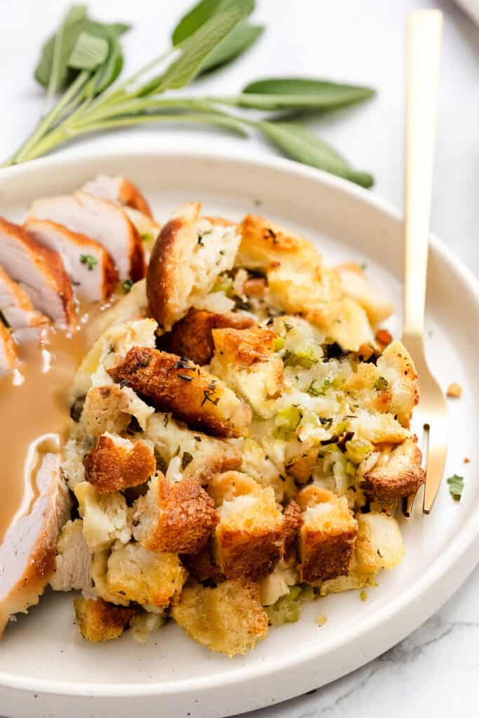 A plate with sliced roasted turkey and make-ahead herb stuffing topped with gravy. A sprig of fresh herbs decorates the edge of the plate, adding a touch of elegance. A gold fork rests beside the food, ready to indulge in this delightful feast.