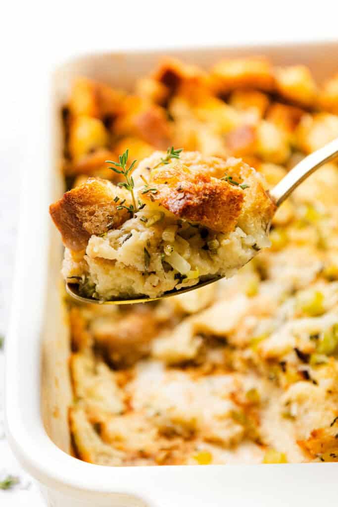 A close-up of a spoonful of herb stuffing over a baking dish. The Thanksgiving side dish is golden brown with pieces of bread, herbs, and vegetables, garnished with a sprig of thyme.